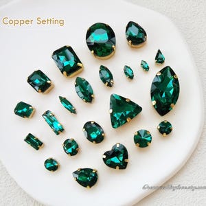 以下が含まれることがあります： 丸型、楕円形、雫型、正方形、長方形など、さまざまな形と大きさのエメラルドグリーンのガラスラインストーンの詰め合わせ。ラインストーンは銅製のセッティングにセットされています。