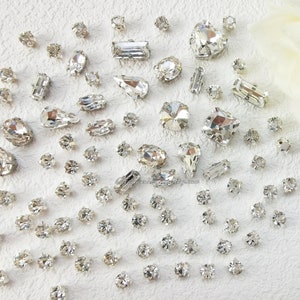 May include: A large assortment of clear crystal rhinestones of various shapes and sizes. The rhinestones are scattered on a white surface.