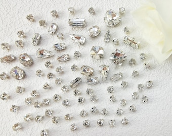 May include: A large assortment of clear crystal rhinestones of various shapes and sizes. The rhinestones are scattered on a white surface.