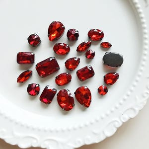 Puede incluir: Una colección de piedras de vidrio rojo facetado en varias formas, incluyendo cortes de pera, ovalados y rectangulares. Cada piedra está montada en un engaste de garras de metal oscuro. Una piedra ovalada se muestra desde atrás, revelando una base de metal oscuro.
