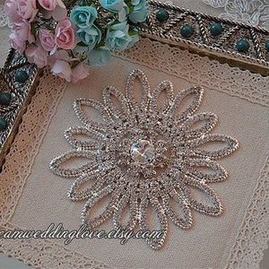 May include: A silver rhinestone embellishment in the shape of a flower with a large clear rhinestone in the center. The embellishment is on a beige fabric background with a lace border.