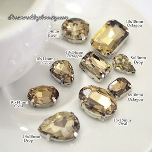 May include: Assortment of clear glass rhinestones in various shapes and sizes, including octagons, ovals, and drops. The rhinestones are set in silver settings and have a light brown color.