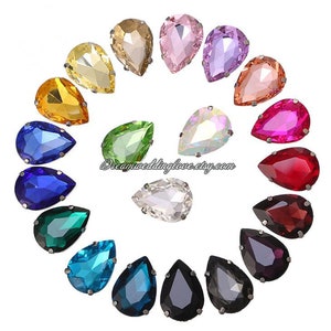 May include: Assortment of 18 teardrop-shaped rhinestones in various colours, including clear, black, blue, green, red, pink, and yellow. The rhinestones are arranged in a circle on a white background.  Dreamweddinglove.etsy.com