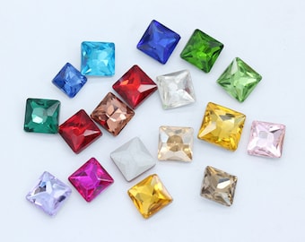 May include: Assortment of square-shaped glass rhinestones in various colours, including red, green, blue, purple, pink, gold, and silver. The rhinestones are arranged on a white surface.