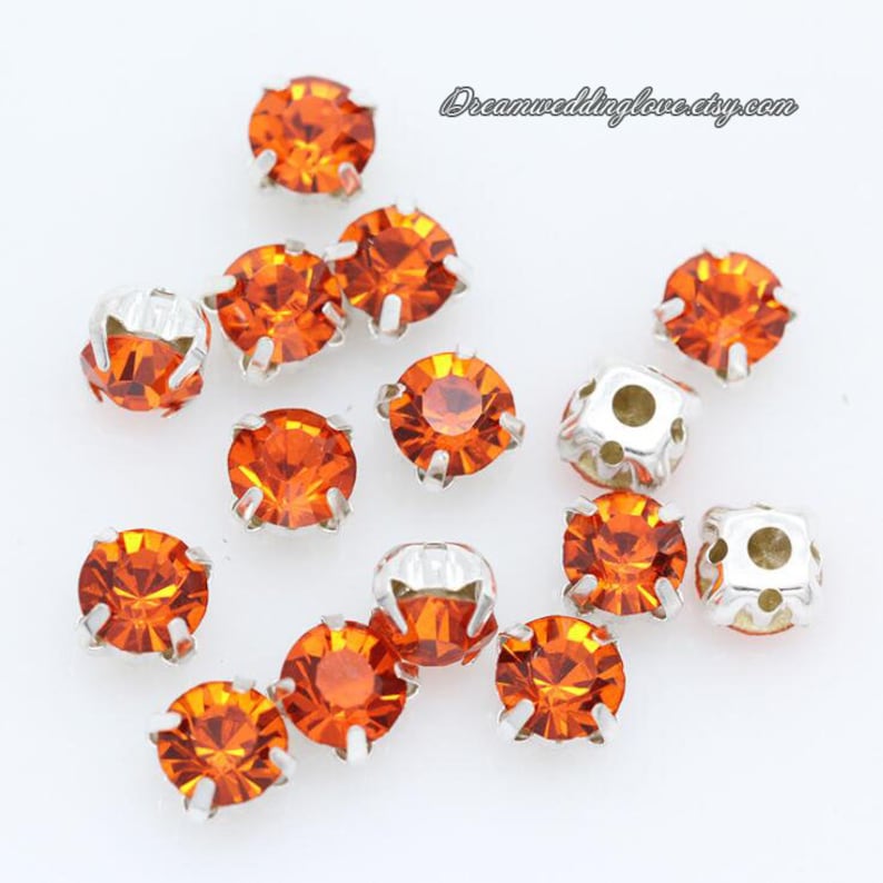 May include: Orange rhinestones with silver settings. The rhinestones are scattered on a white surface. Dreamweddinglove.etsy.com is visible in the background.