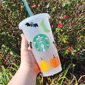 Peut inclure: Un gobelet réutilisable en plastique transparent avec une paille verte. Le gobelet est décoré d'autocollants sur le thème d'Halloween, notamment une chauve-souris, un croissant de lune, une citrouille et une sorcière. Le gobelet est tenu par une main.