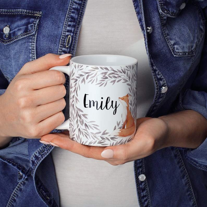 May include: White ceramic mug with a grey and white leafy design and a fox illustration. The mug has the name "Emily" written in black lettering.