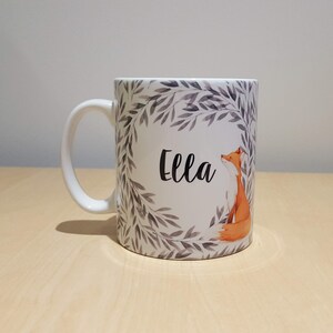 May include: White ceramic mug with a grey and white leafy design and a fox illustration. The mug has the name "Ella" written in black cursive lettering.