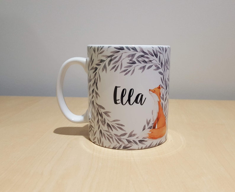 May include: White ceramic mug with a grey and white leafy design and a fox illustration. The mug has the name "Ella" written in black cursive lettering.