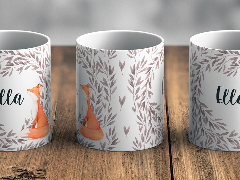 May include: Two white ceramic mugs featuring a watercolour design of grey leaves and a fox. The mugs are personalised with the name "Ella" printed on them.