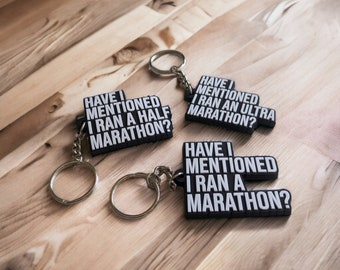 Puede incluir: Tres llaveros negros con texto blanco. Cada llavero dice "Have I mentioned I ran a marathon?" con diferentes variaciones de la distancia del maratón. Los llaveros están sobre una superficie de madera.