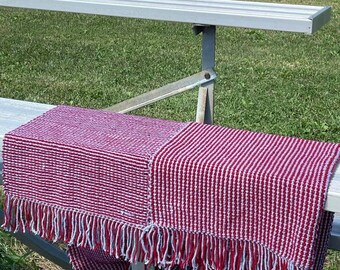 May include: A red and white woven throw blanket with fringe is draped over a metal bench. The blanket has a striped pattern and is made of a textured material. The background shows a grassy park with trees.