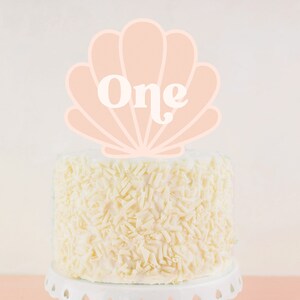 May include: A white cake with white chocolate shavings on top, decorated with a pink seashell cake topper that says "One" in white letters.