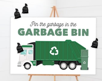 May include: A white sign on an easel with the text "Pin the garbage in the GARBAGE BIN." The sign features a green garbage truck with a recycling symbol and a green trash can. Black garbage bags are pinned around the sign.