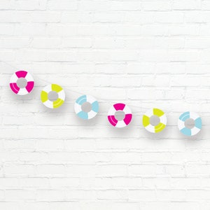 Puede incluir: Una pared de ladrillo blanco con una cadena de decoraciones de papel con forma de salvavidas de colores que cuelgan de ella. Las decoraciones son en tonos de rosa, amarillo y azul.