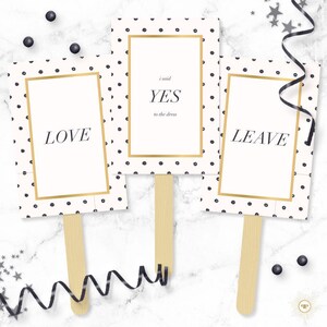 Op de afbeelding: Drie witte kartonnen borden met zwarte stippen en gouden randjes op houten stokjes. De borden zeggen "LOVE", "Ik zei JA tegen de jurk" en "LEAVE".