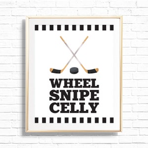 May include: A framed print with a white background and black text that reads "WHEEL SNIPE CELLY". Two hockey sticks are crossed in the center of the print with a black hockey puck underneath. The top and bottom of the print have a black and white striped border.