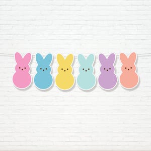 May include: A decorative Easter banner featuring six bunny shapes in pastel colours: pink, blue, yellow, light blue, purple, and peach. Each bunny has a white border and three black dot eyes. The banner is strung on a thin white string against a white brick wall.