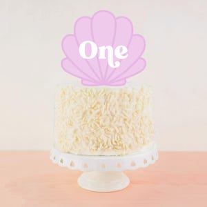 May include: A white cake with white sprinkles topped with a pink seashell cake topper that says "One" in white letters.