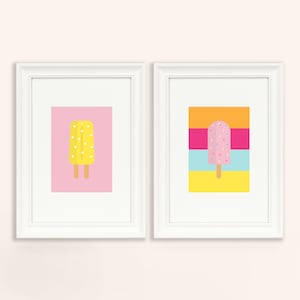 May include: Two framed prints of ice lollies. The first print features a yellow ice lolly with white dots on a pink background. The second print features a pink ice lolly with colourful sprinkles on a background of orange, pink, blue, and yellow stripes.