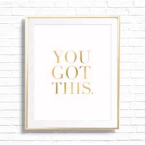 May include: A framed print with the words "YOU GOT THIS." in gold lettering on a cream background. The frame is a light gold color, and the background is a white brick wall.