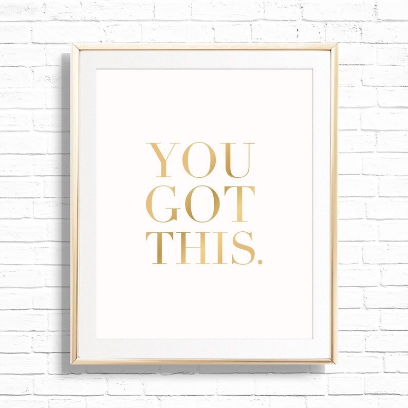 May include: A framed print with the words "YOU GOT THIS." in gold lettering on a cream background. The frame is a light gold color, and the background is a white brick wall.