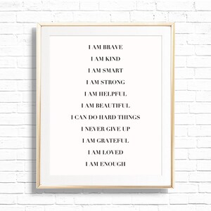 Op de afbeelding: Een ingelijste print met een witte achtergrond en zwarte tekst. De tekst bevat affirmaties zoals "IK BEN MOEDIG", "IK BEN LIEF" en "IK BEN GENOEG". De lijst is licht van kleur en de achtergrond is een witte bakstenen muur.