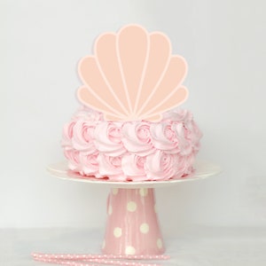 May include: A pink cake with a seashell cake topper, presented on a white cake stand with a pink polka-dot base. Pink paper straws are in the foreground. The cake is decorated with pink frosting roses.