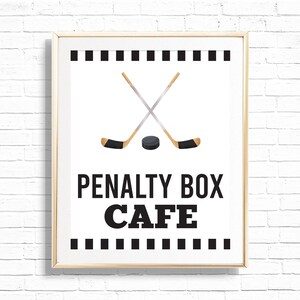 Può includere: Un cartello bianco con bordo nero e testo nero che recita "Penalty Box Cafe". Il cartello presenta una coppia di bastoni da hockey incrociati e un disco da hockey nero.