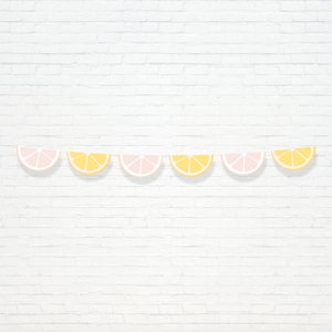 May include: A decorative banner with alternating pink and yellow citrus fruit slices. The banner is strung against a white brick wall, creating a cheerful, summery aesthetic. The fruit slices are evenly spaced, adding a pop of colour.