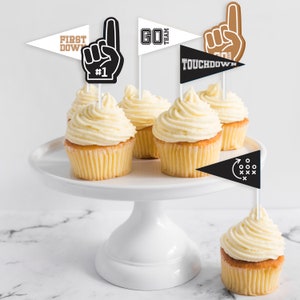 Può includere: Un porta torte bianco con cinque cupcake decorati con glassa bianca. I cupcake sono sormontati da vari topper a tema calcistico, tra cui una mano bianca e nera con il numero "#1", un pennant bianco e nero con la parola "GO" e la parola "TEAM", un pennant bianco e nero con la parola "TOUCHDOWN", e un pennant bianco e nero con un pallone da calcio e le parole "FIRST DOWN".