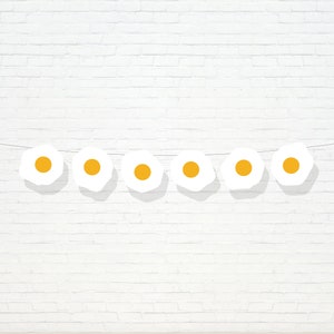 May include: A string of white paper eggs with yellow yolks hanging against a white brick wall.