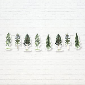 May include: A garland of nine watercolor pine trees, each with a white outline, strung together with white string. The trees are green and brown and are arranged in a row against a white brick wall.