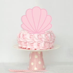 Op de afbeelding: Een roze cake met een zeeschelp taarttopper, gepresenteerd op een witte taartstandaard met een roze stippenbasis. Roze en wit gestreepte rietjes staan onderaan de afbeelding. De taart is versierd met roze rozen van glazuur.