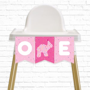 May include: Pink high chair banner with the word "ONE" in white letters. The middle letter "N" is replaced with a pink frosted animal cookie wearing a party hat. The banner has a pink background with white and multi-colored confetti.