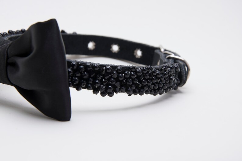 May include: Black dog collar with a satin bow and a beaded section. The collar is made of black leather and has a silver-colored buckle and eyelets. The beaded section is made of small, black beads.