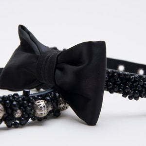 May include: Black dog collar with a satin bow tie. The collar is adorned with black beads and silver accents. The leather collar has blue stitching.