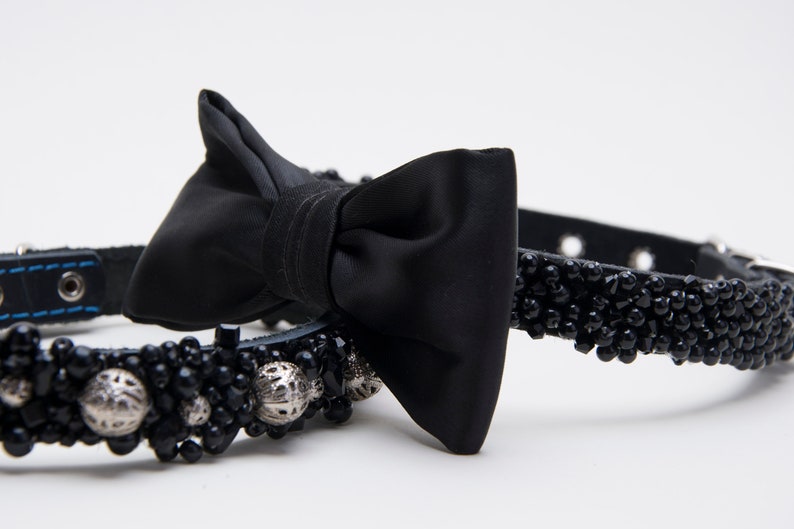 May include: Black dog collar with a satin bow tie. The collar is adorned with black beads and silver accents. The leather collar has blue stitching.