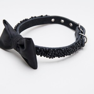 May include: Black dog collar with a bow tie. The collar is made of black leather and is adorned with small black beads. The bow tie is made of a smooth, black fabric. The collar has a metal buckle and a metal loop.