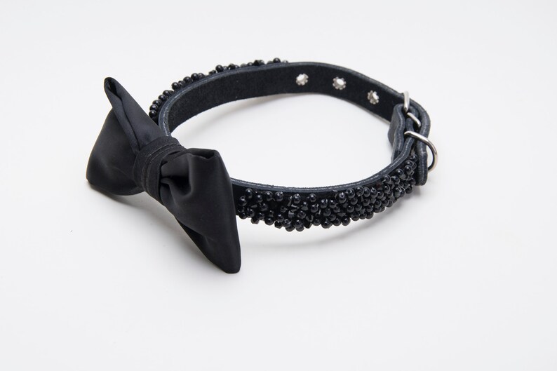 May include: Black dog collar with a bow tie. The collar is made of black leather and is adorned with small black beads. The bow tie is made of a smooth, black fabric. The collar has a metal buckle and a metal loop.