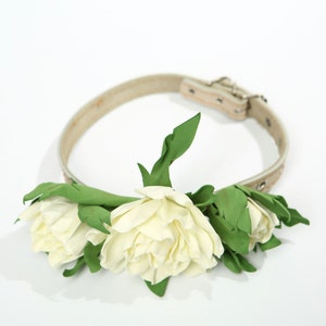 Puede incluir: Un collar de perro de cuero blanco con una hebilla plateada, decorado con tres flores de tela blanca y hojas verdes.
