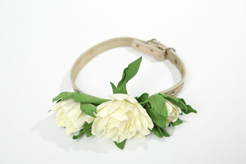 Puede incluir: Un collar de perro de cuero blanco con una hebilla plateada, decorado con tres flores de tela blanca y hojas verdes.