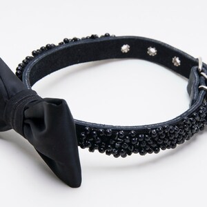 May include: Black dog collar with a satin bow tie. The collar is made of black leather and is embellished with small, black beads. The collar has a metal buckle and several holes for adjustment. A stylish accessory for pets.