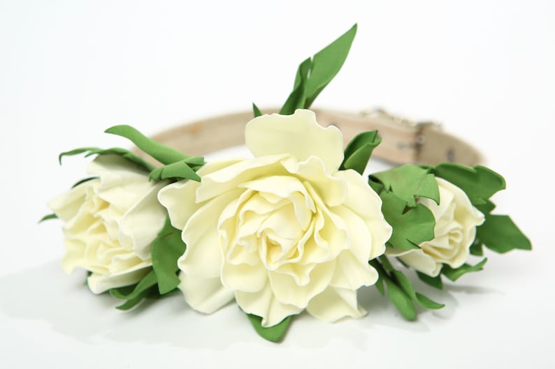 Puede incluir: Un collar de perro de cuero blanco con tres flores de tela blancas y hojas verdes.