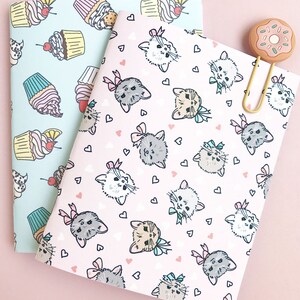 May include: Two notebooks with different patterns. One notebook has a light blue background with a repeating pattern of cupcakes. The other notebook has a pink background with a repeating pattern of kittens with bows. A gold paperclip with a pink donut charm is attached to the notebooks.