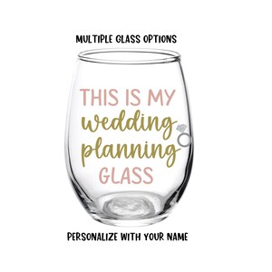 May include: A clear glass wine glass with the text "This is my wedding planning glass" in gold and pink lettering. A small silver ring charm is attached to the glass.