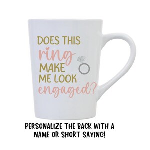 May include: White ceramic coffee mug with a pink and gold design that says "Does this ring make me look engaged?"  The mug has a handle and is personalized with the text "Personalize the back with a name or short saying!"