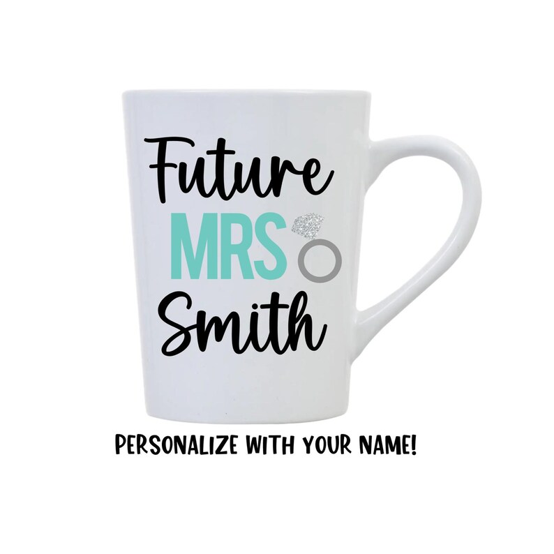 K&ouml;nnte beinhalten: Wei&szlig;e Keramiktasse mit schwarzem Text, der "Future Mrs. Smith" lautet, mit einem t&uuml;rkisfarbenen "Mrs." und einem silbernen Ring mit einem Diamanten. Der Text "Personalisieren Sie mit Ihrem Namen!" befindet sich unter der Tasse.