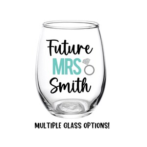 May include: A clear glass wine glass with the text "Future Mrs. Smith" in black lettering. A turquoise blue "MRS" is centered on the glass with a silver glitter ring above it.