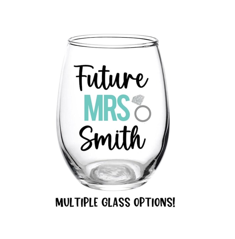 May include: A clear glass wine glass with the text "Future Mrs. Smith" in black lettering. A turquoise blue "MRS" is centered on the glass with a silver glitter ring above it.
